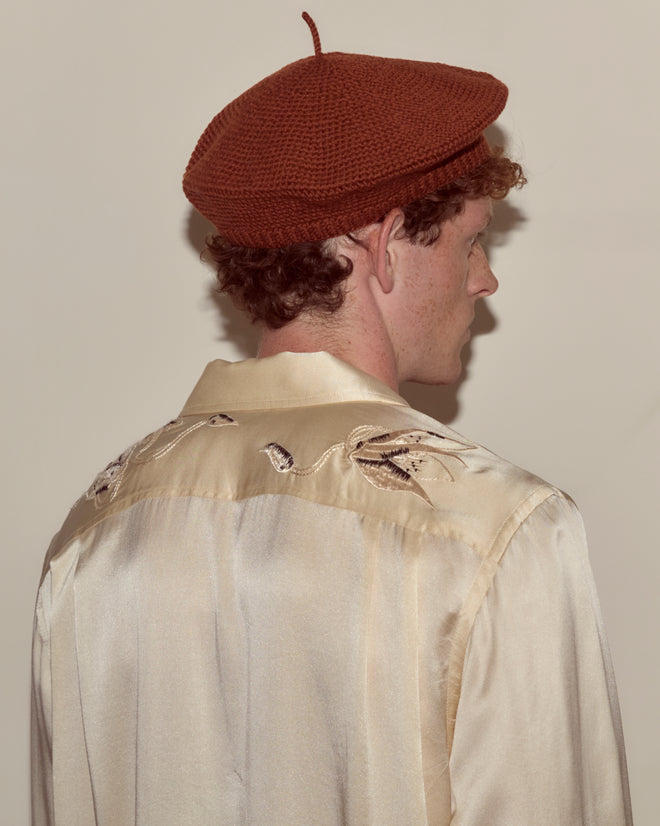 A person in a tobacco-colored crochet beret and a silk shirt with embroidered details, facing away from the camera.