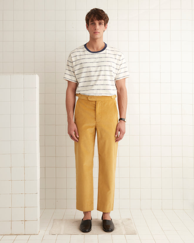 A pair of Custom Senior Cord Side-Buckle Trousers in light brown, featuring pockets, is hanging on a wooden hanger against a white wall with hooks.