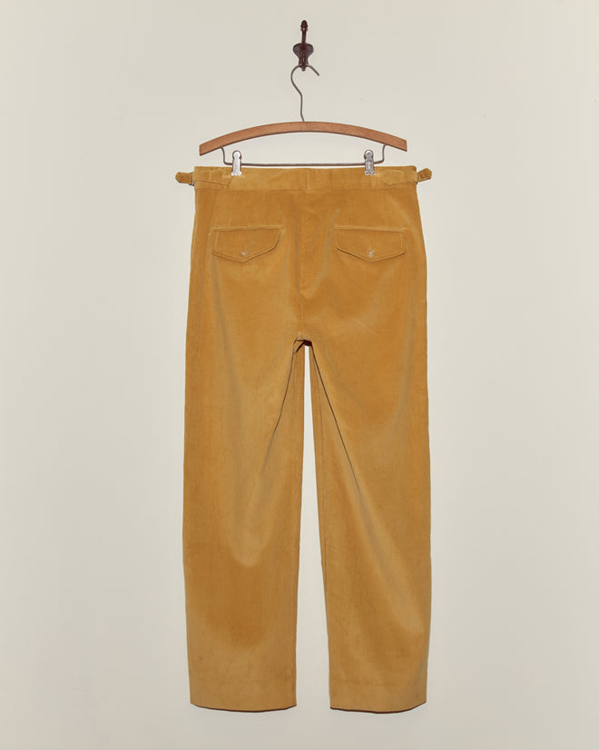 Custom Senior Cord Side-Buckle Trousers in mustard yellow corduroy hang on a wooden hanger against a plain white wall.