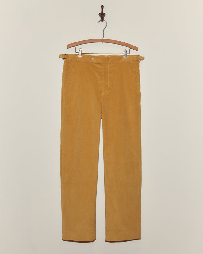 Custom Senior Cord Side-Buckle Trousers in mustard yellow hang on a wooden hanger against an off-white wall, highlighting classic British corduroy style.