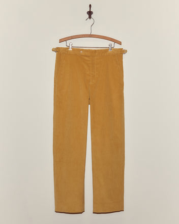 Custom Senior Cord Side-Buckle Trousers in mustard yellow hang on a wooden hanger against an off-white wall, highlighting classic British corduroy style.