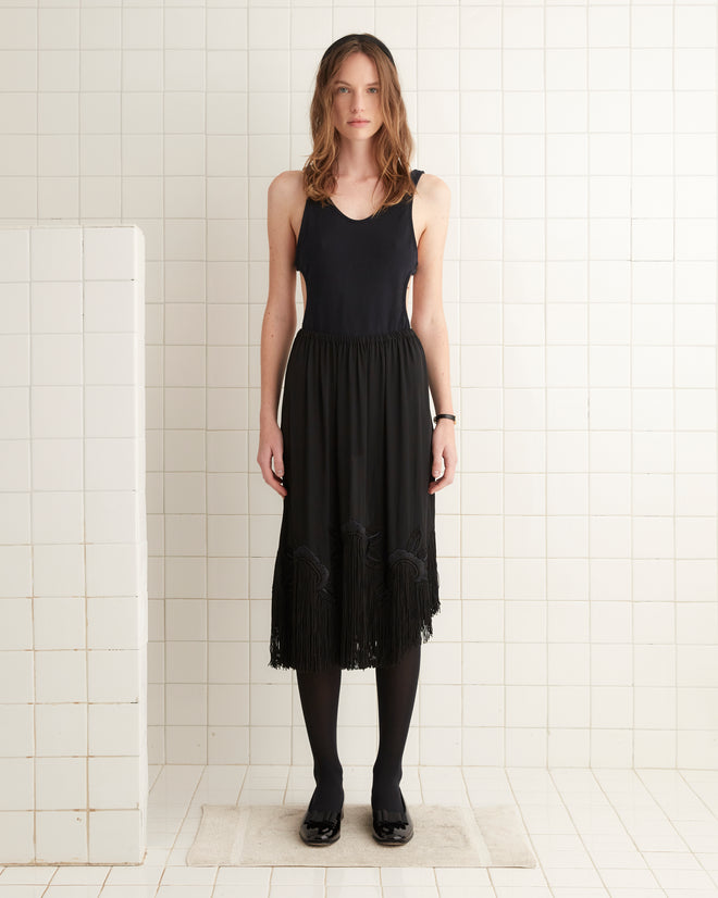 A woman in a Daffodil Fringe Skirt, featuring satin-stitched floral embroidery, stands in a tiled room wearing tights, her expression neutral.