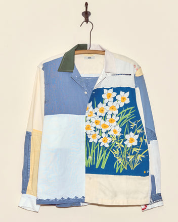 The Daffodil Napkin Shirt - S/M, featuring a vintage floral patchwork design, hangs on a hook against a plain light wall.
