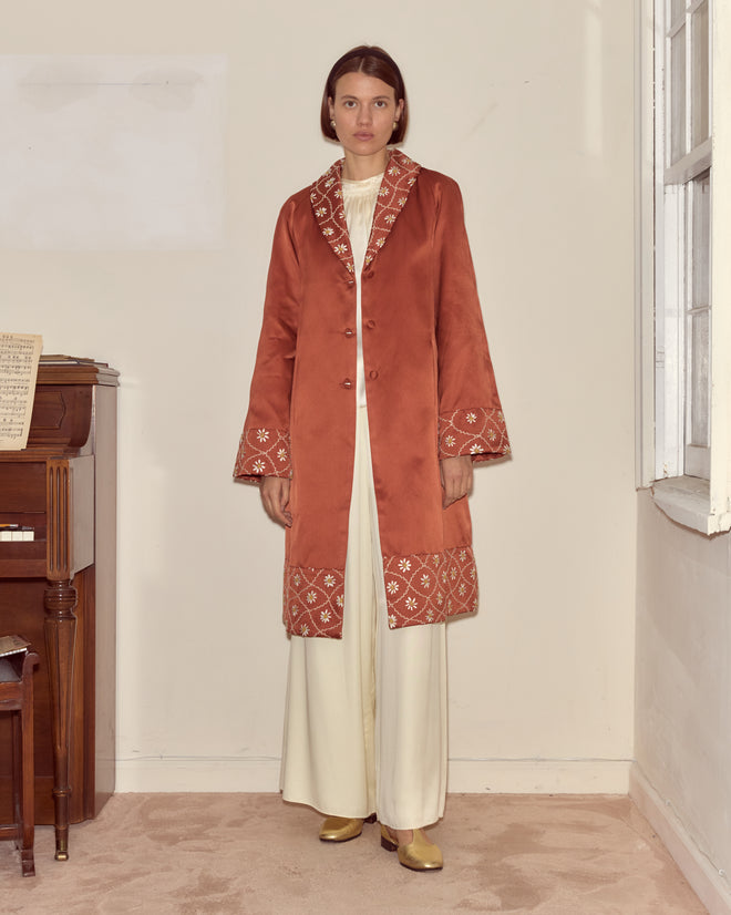 A woman stands indoors by a piano and window, facing forward, wearing the Daisy Chain Coat—a long rust-colored 1950s style with embroidered trim and a couture silhouette.