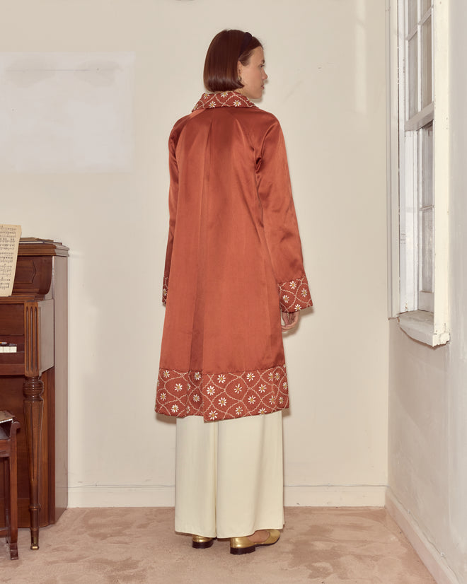 A woman wearing the Daisy Chain Coat—an elegant, long rust-colored 1950s design with white embroidery—stands indoors, back to the camera, facing a window and highlighting the couture silhouette.
