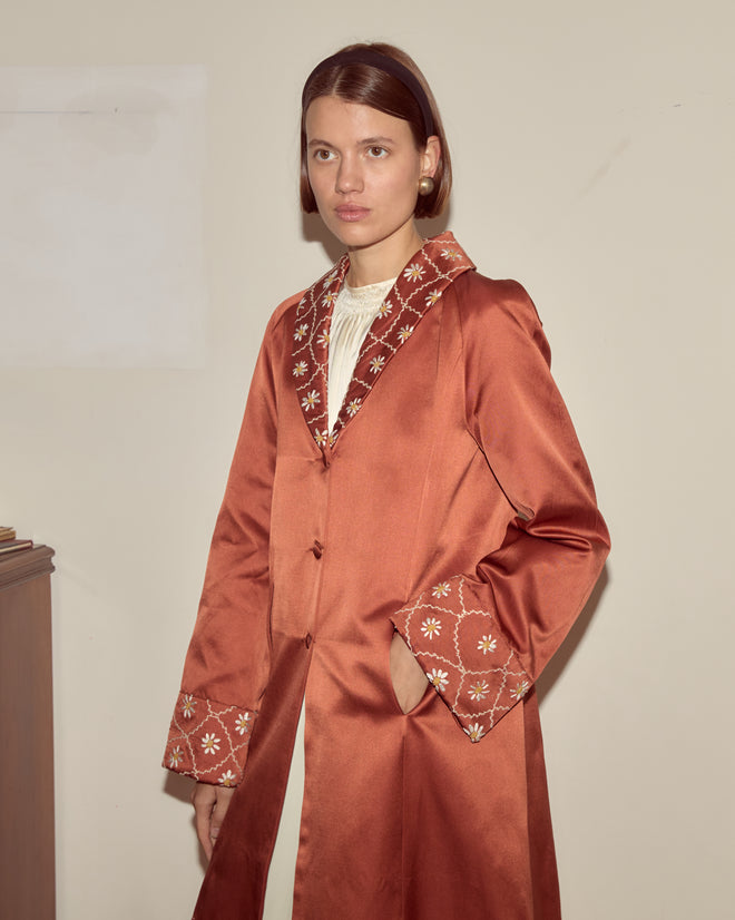 Woman indoors against a plain wall wears the Daisy Chain Coat, a rust-colored 1950s-inspired piece featuring embroidered details and an antique daisy pattern.