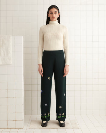 The Daisy Garden Joggers - Green with a beadwork floral motif are hanging on a wooden hanger against a white tile wall.