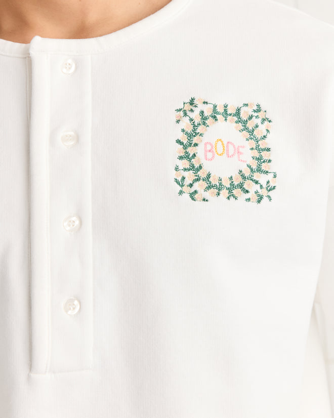 Close-up of the Daisy Never Tell Henley Sweatshirt, a 100% cotton white top with buttons and colorful "BODE" embroidery, surrounded by green leaves.