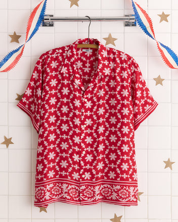 The Cherry Daisy Short Sleeve Shirt, featuring red silk with a white floral pattern inspired by vintage bandana prints and Indian textiles, hangs on a hanger against a tiled wall adorned with star and ribbon decor.