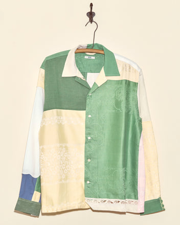 The Damask Greens Napkin Shirt (L/XL) features unique green, yellow, blue, and cream patchwork panels. Handmade in New York, it hangs boldly on a wall hook—a true one-of-a-kind vintage piece.