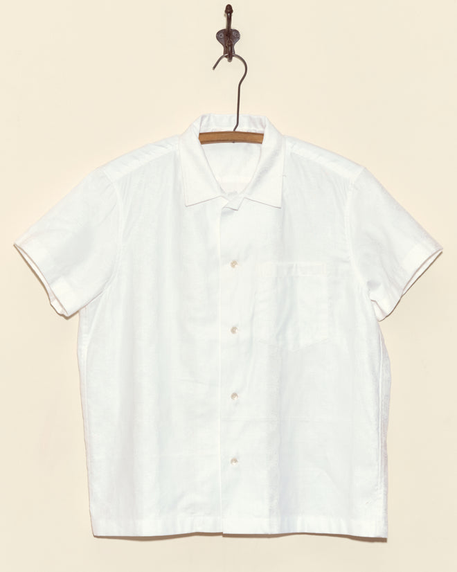 The Damask Moonflower Short Sleeve shirt, a white button-up with short sleeves, hangs on a hanger against a beige wall.
