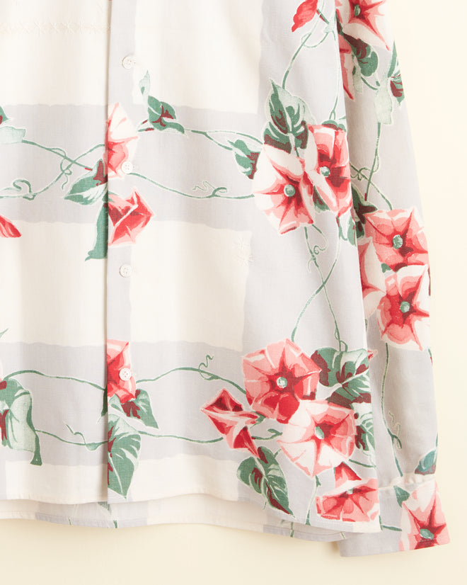 The Dandy Dianthus Shirt (Size S/M), adorned with colorful florals in shades of red and pink on a white background reminiscent of a midcentury tablecloth, is elegantly displayed hanging on a wall hook.