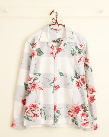 The Dandy Dianthus Shirt (Size S/M), adorned with colorful florals in shades of red and pink on a white background reminiscent of a midcentury tablecloth, is elegantly displayed hanging on a wall hook.