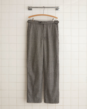 The Dark Grid Trousers - 31, crafted from 100% wool, hang elegantly on a wooden hanger against a tiled wall, showcasing their black and white checkered pattern and unique charm.