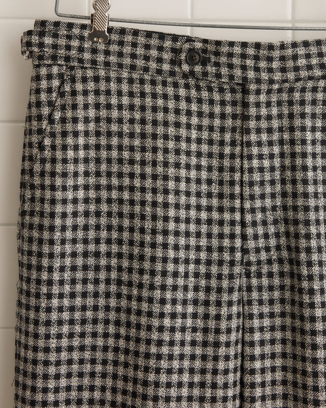 The Dark Grid Trousers - 31, crafted from 100% wool, hang elegantly on a wooden hanger against a tiled wall, showcasing their black and white checkered pattern and unique charm.