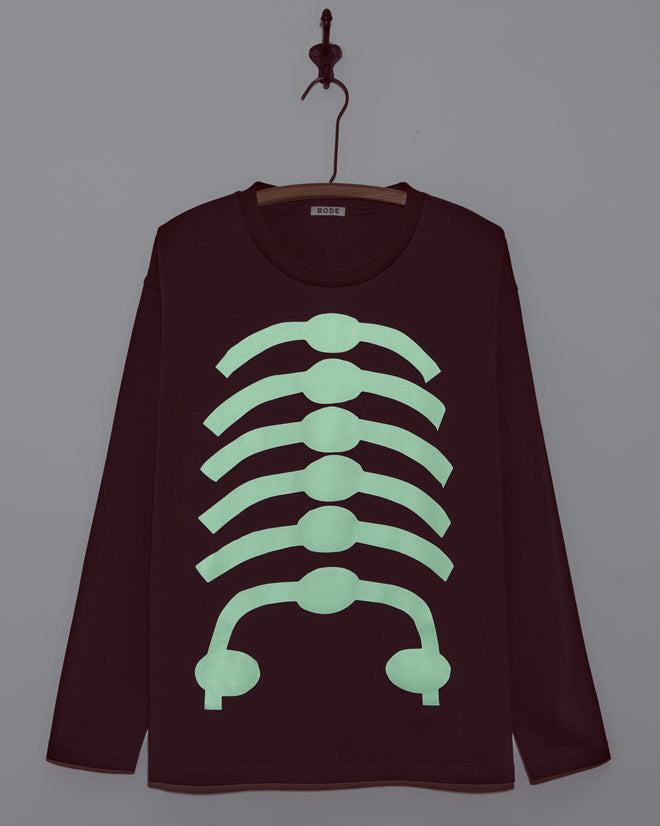 The Skeleton Long Sleeve Tee is a black boxy-fit shirt featuring a green glow-in-the-dark ribcage design, displayed on a hook against a gray wall.