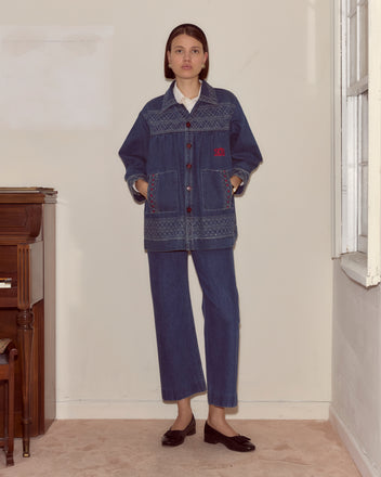 A woman stands indoors by a window, wearing an Embroidered Denim Quincy Jacket, matching denim pants, a white shirt, and black shoes.