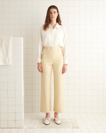 A person stands in a tiled room wearing a white blouse, Dennis Stripe Sailor Trousers with antique pearl buttons, and white loafers.