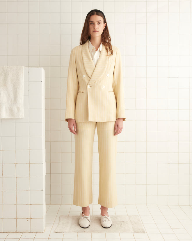 A woman stands in front of a tiled wall wearing the Dennis Stripe Suit Jacket, which features a cream-colored pinstripe design and antique pearl buttons, paired with white shoes. A towel hangs on a tiled surface to her left.