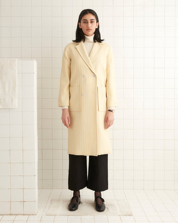 A woman in a Dennis Stripe Worcester coat, featuring a vintage cream double-breasted design with pearl buttons, stands elegantly in a tiled room, complemented by her black pants and matching shoes.