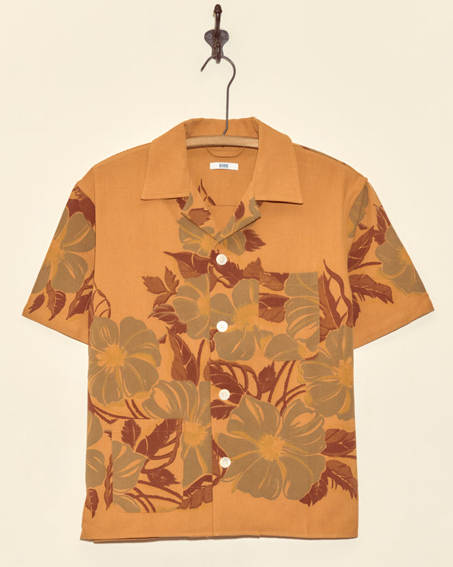 The Desert Flower Short Sleeve Shirt - XS/S features a unique brown and mustard floral print, displayed hanging on a hook against a plain wall for an elegant touch.