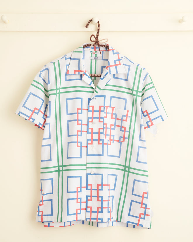 Dessau Maze Shirt - XS/S featuring short sleeves and a vibrant geometric pattern of blue, green, and red squares, hanging on a white wall hook.