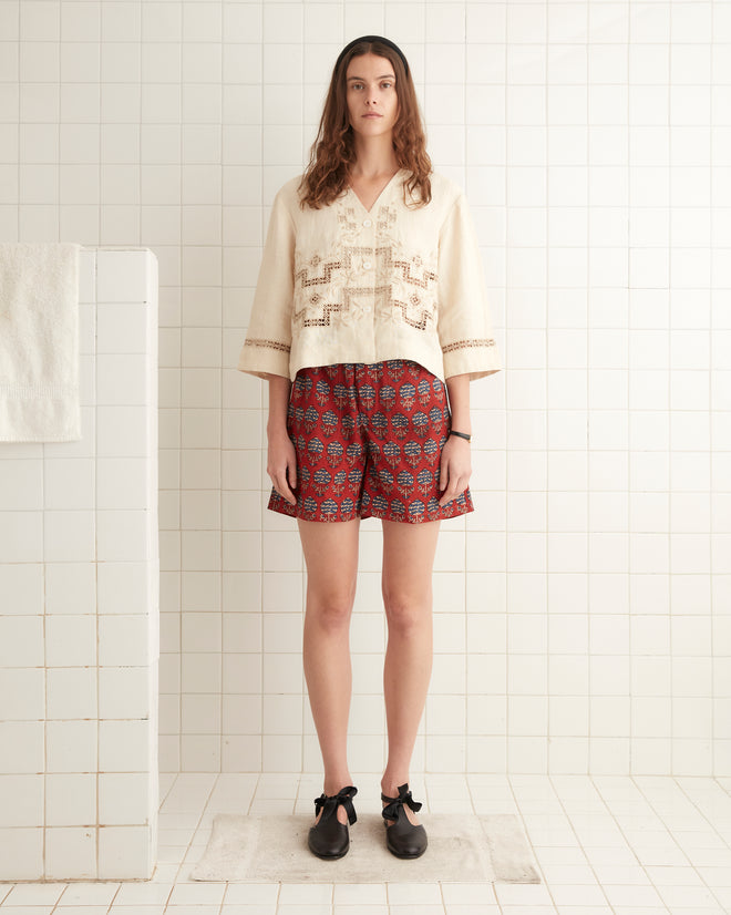 A person with long hair stands in a tiled bathroom, wearing a beige blouse with embroidery and Dahlia Block Print Rugby Shorts made in India. They wear black shoes with bows, and a white towel hangs nearby.