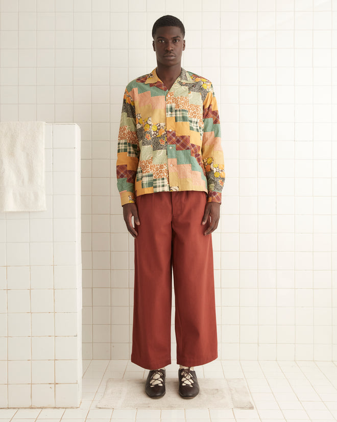 A person stands against a tiled wall wearing a Diagonal Square Patchwork Shirt and loose-fitting red pants. A white towel hangs on a nearby wall.