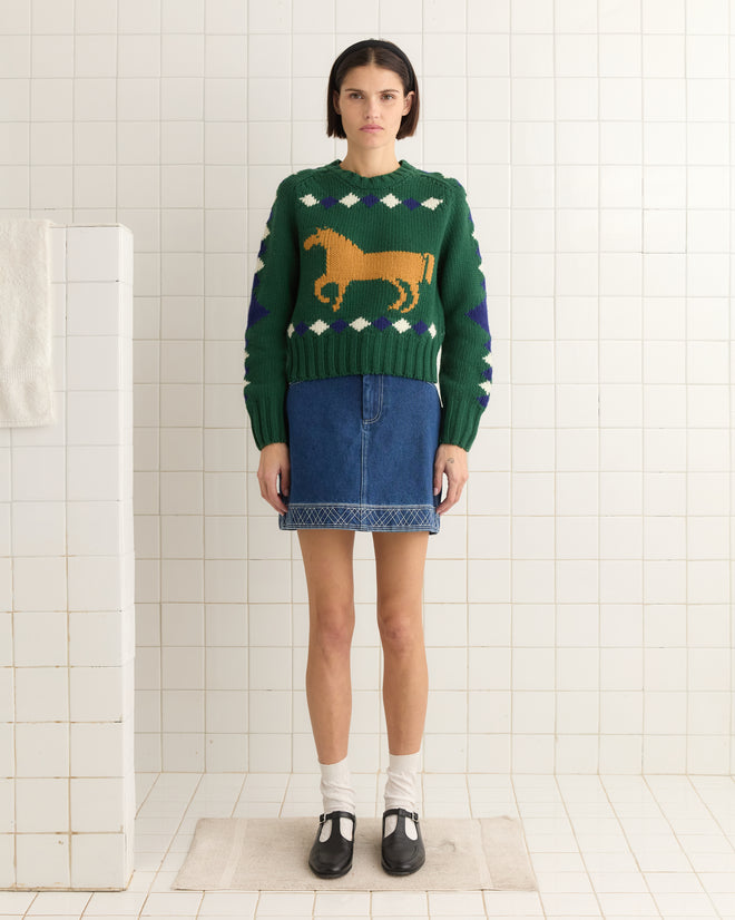 In a tiled room, a person wears the green Diamond Corral Sweater with a horse design, paired with a denim skirt and black shoes.