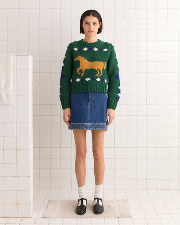 A person in a green Diamond Corral Sweater with a horse pattern and a denim skirt stands in a tiled room.