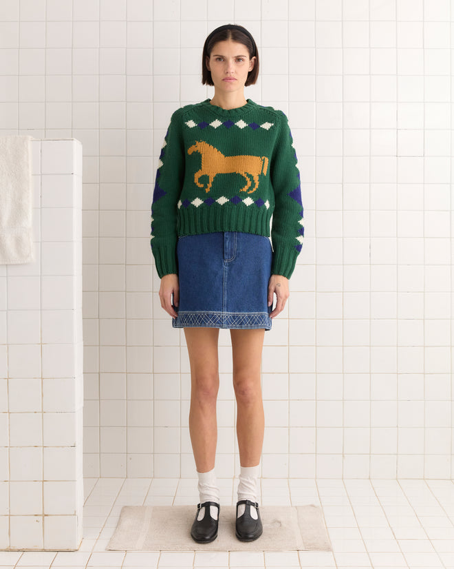 A person in a green Diamond Corral Sweater with a horse pattern and a denim skirt stands in a tiled room.