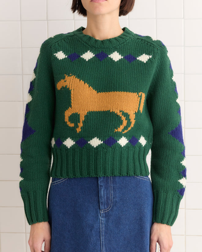 A person wears the Diamond Corral Sweater, a green merino wool piece featuring a brown horse design and geometric patterns, paired with a blue skirt.