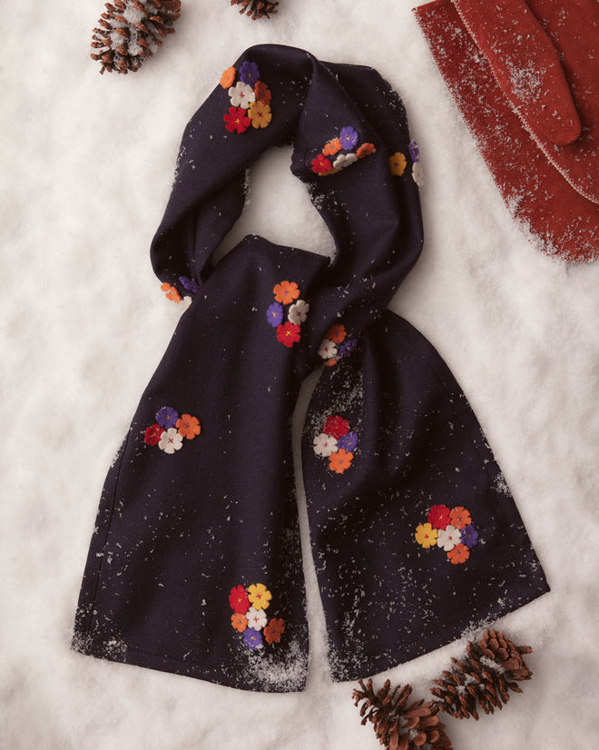 The Dianthus Appliqué Scarf, crafted from 100% wool in navy and embellished with vibrant felt flowers, rests on the snow, surrounded by pine cones and a nearby red glove.