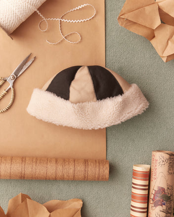 The Patchwork Shearling Hat, inspired by the 1960s, is stylishly positioned on a green surface, with scissors, brown paper, and ribbon surrounding it.