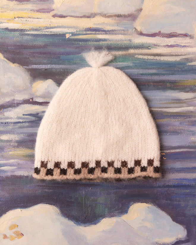 The Edge Check Hat, featuring a delicate jacquard-knit design with white and brown checkered trim, is set against a painted backdrop of clouds and sky, evoking a sense of timeless elegance.