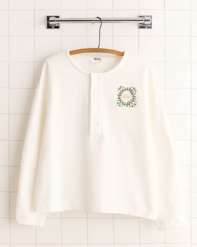 The Daisy Never Tell Henley Sweatshirt is a long-sleeve, 100% cotton white top with "BODE" and a green floral wreath embroidered on the chest. It features a boxy fit for a relaxed look.