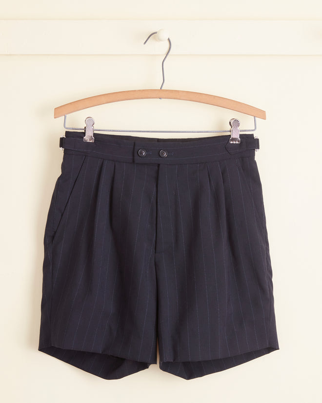 A pair of Digit Stripe Wool Shorts - 27 in black deadstock wool with pinstripes, featuring pleats, button closure, and side pockets, is hanging on a wooden hanger against a light-colored wall.