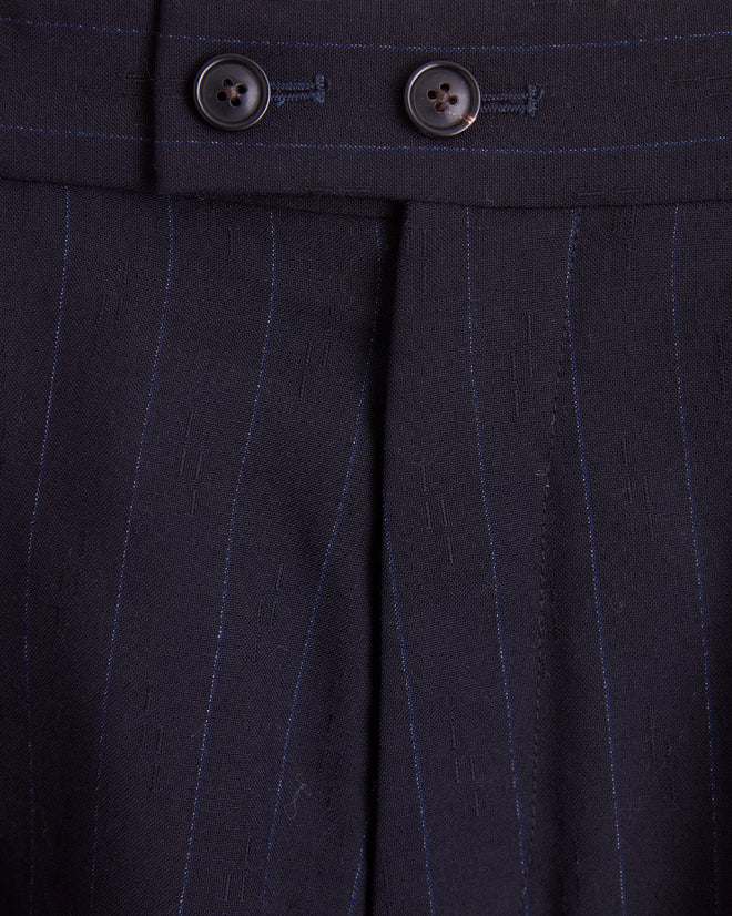 A pair of Digit Stripe Wool Shorts - 27 in black deadstock wool with pinstripes, featuring pleats, button closure, and side pockets, is hanging on a wooden hanger against a light-colored wall.