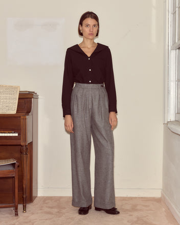 A woman stands indoors by a piano, wearing a black top and the Dovecote Port Trousers—classic heathered grey suiting crafted from wool by a renowned UK mill.