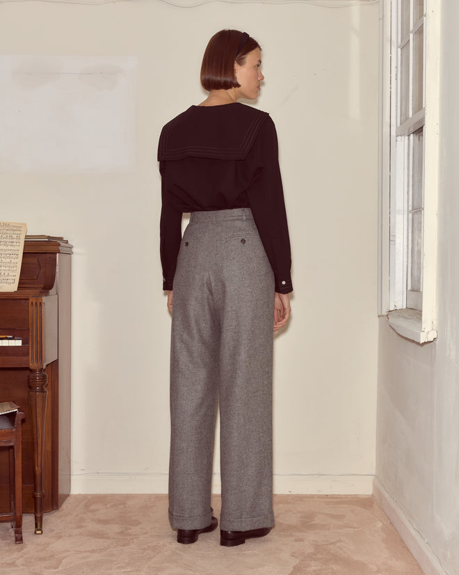 A woman with short brown hair in a black top and classic suiting—Dovecote Port Trousers—stands facing a wall near a piano and window.