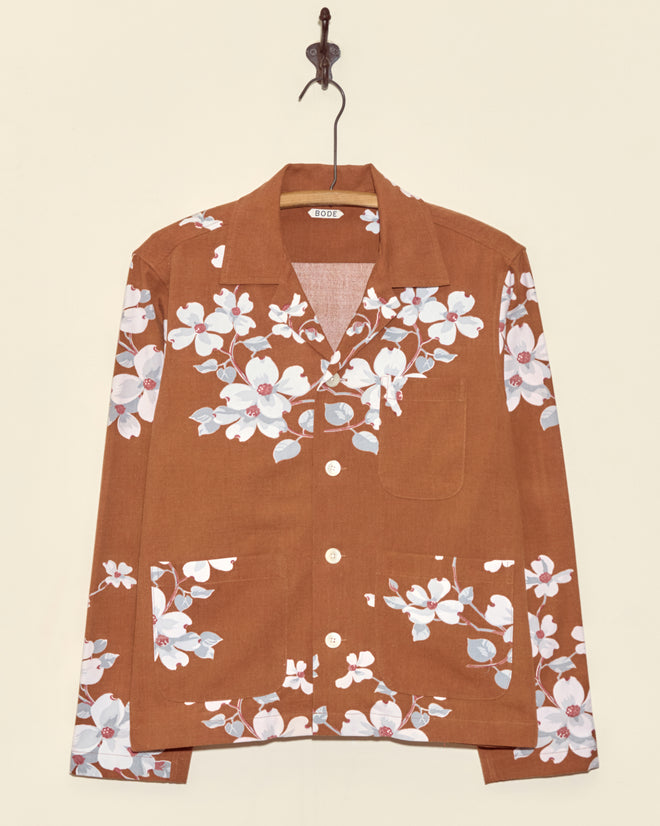 The Dogwood Bark Long Sleeve Shirt - XS is a unique brown button-up with a white and pink floral print, shown hanging on a hook against a beige wall.