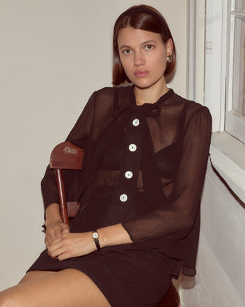 A woman in the Dottie Blouse, featuring sheer silk fabric and antique pearl buttons, sits by a window on a chair, looking at the camera.