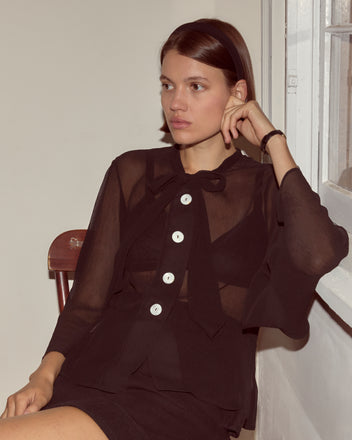 A woman in the Dottie Blouse, a sheer silk top with antique pearl buttons, sits thoughtfully on a wooden chair by the window.