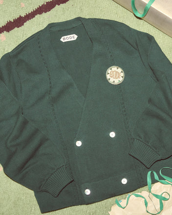 The Double-Breasted Cardigan in dark green by Bode, featuring white buttons, is displayed on a green textured surface.