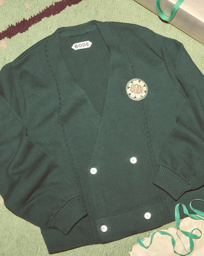 The Double-Breasted Cardigan in dark green by Bode, featuring white buttons, is displayed on a green textured surface.