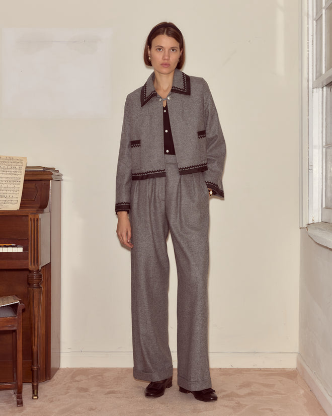 A woman wearing the Dovecote Trim Jacket in heathered grey with silver ship buttons stands indoors by a piano with sheet music near a window.