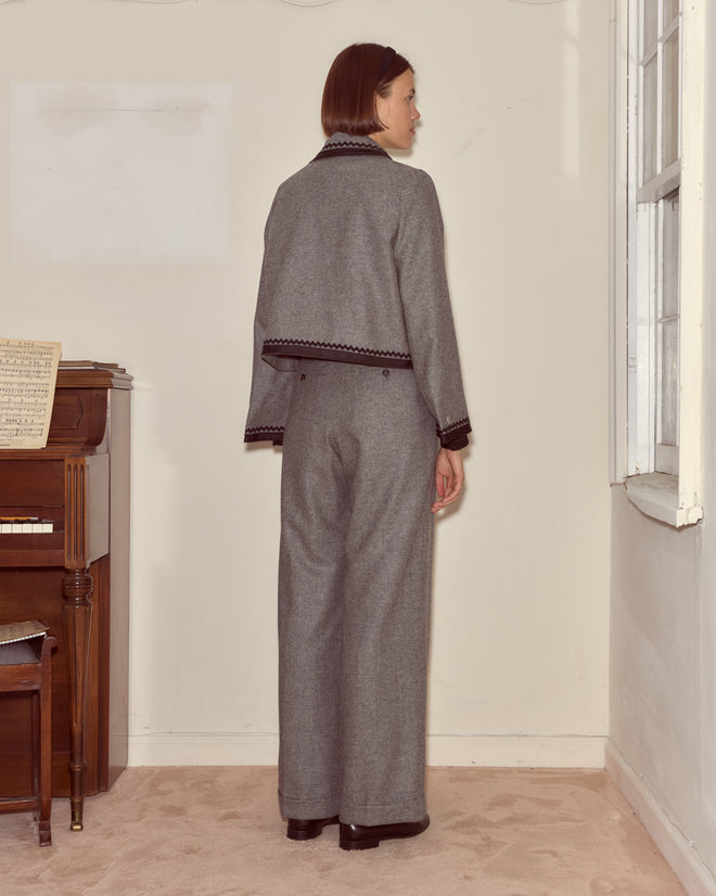 A person wearing the Dovecote Trim Jacket in heathered grey stands with their back to the camera near a piano and a window in a beige room.