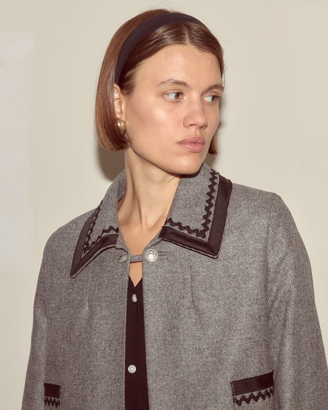 Woman with short brown hair and headband wearing the Dovecote Trim Jacket, a heathered grey wool style with black trim and silver ship buttons, looking to the side.