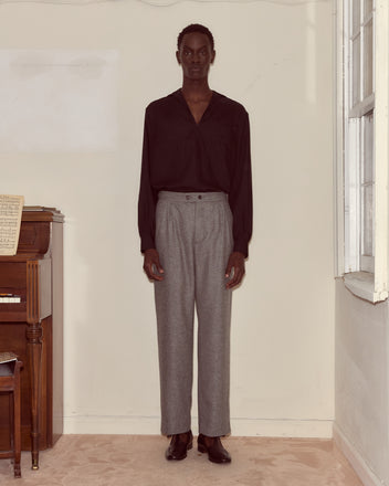 A person stands indoors by a piano, dressed in a black shirt and classic Dovecote Trousers, gazing directly ahead.