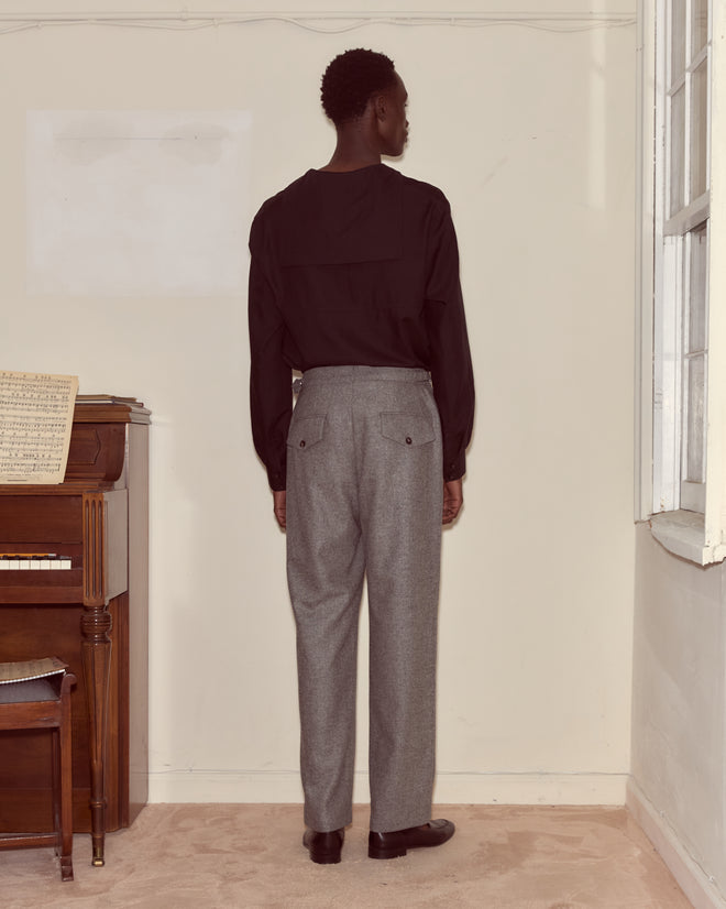 A person in a black shirt and classic Dovecote Trousers stands facing a wall near a piano in a softly lit room.